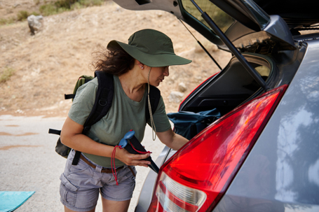 A woman wearing a hat and outdoor clothing organizes her belongings from a car trunk, preparing for a hiking or camping trip in a warm, natural setting.