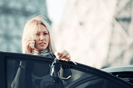 Woman with blonde hair standing next her car door, talking on cell phone